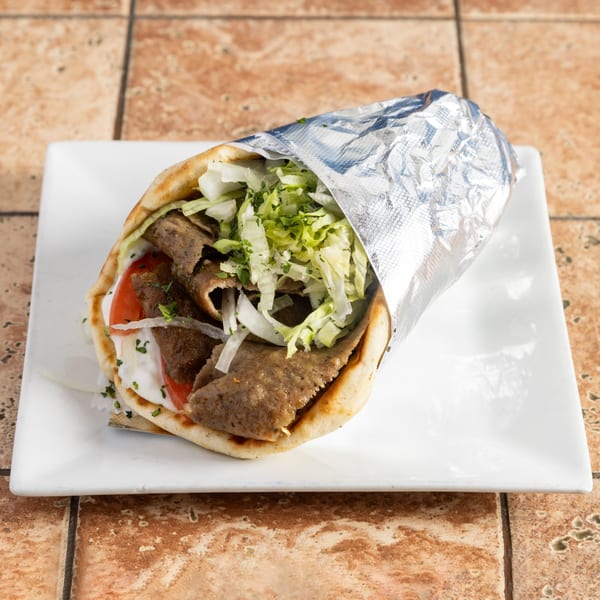 A gyro sandwich with meat, lettuce, tomatoes, onions, and white sauce, partially wrapped in foil on a white plate.