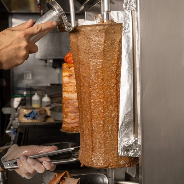A chef's hand using a metal shaver to slice meat from a large vertical rotisserie.
