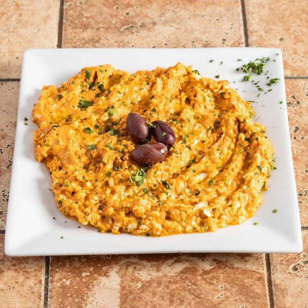 A white rectangular plate holds a vibrant orange dip, garnished with parsley and three black olives.