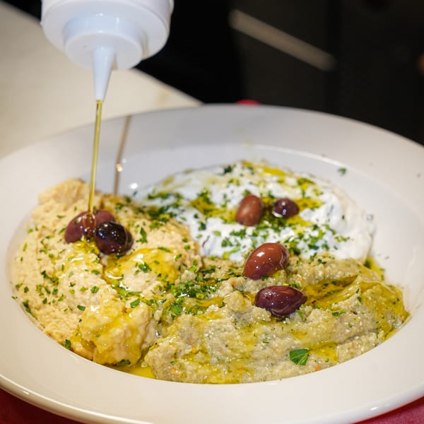 Olive oil drizzling over a white bowl of hummus, baba ghanoush, and tzatziki with olives.