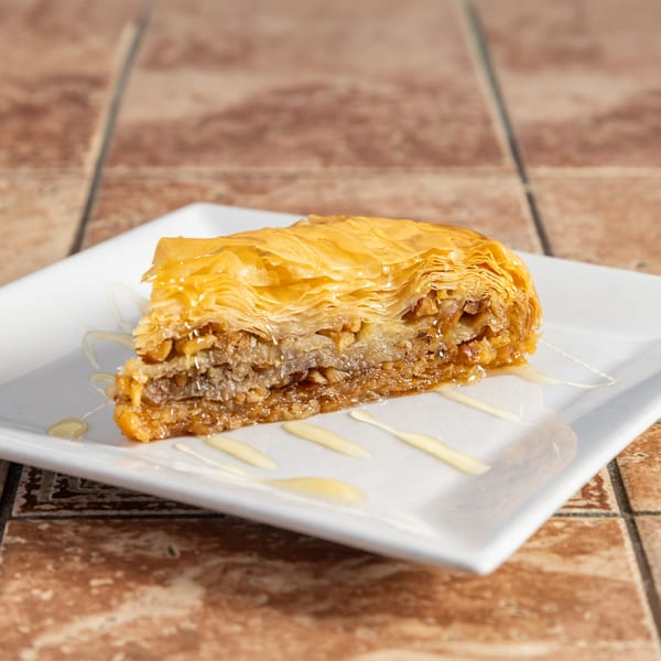 A triangular slice of golden-brown baklava, drizzled with honey, on a white rectangular plate.