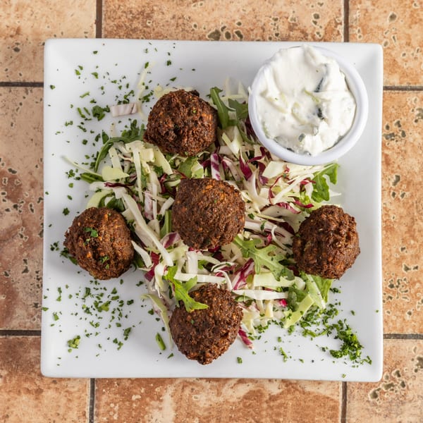 Five falafel balls on a bed of shredded cabbage and arugula, with a side of white sauce.